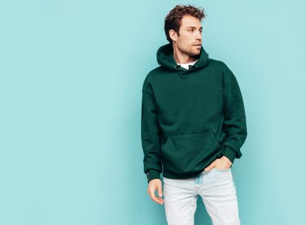 Handsome confident hipster  model.Sexy unshaven man dressed in summer stylish green hoodie and jeans clothes. Fashion male with curly hairstyle posing in studio. Isolated on blue